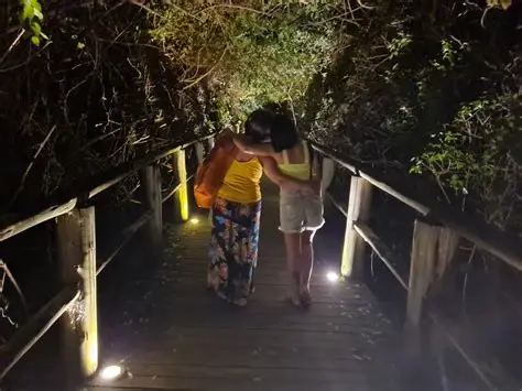 Duas mulheres abraçadas caminhando por uma passarela de madeira iluminada à noite, em meio à vegetação