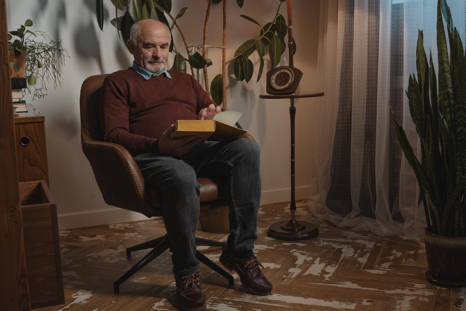 Homem idoso lendo um livro sentado em uma poltrona, em uma sala aconchegante com plantas